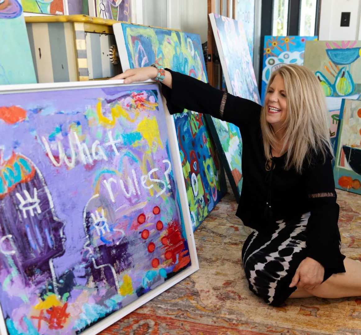 Artist Aleea Jaques smiles as she sits next to a purple abstract painting with the words "What rules?" painted on the canvas.