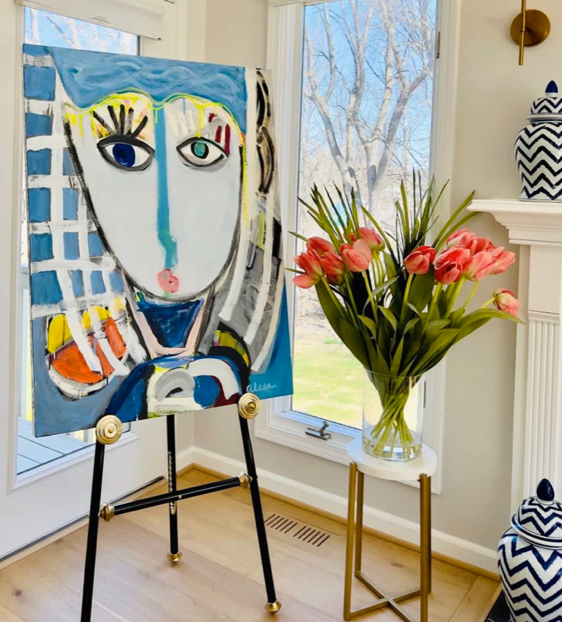 A blue abstract portrait painting sits on a display easel in a contemporary living room.
