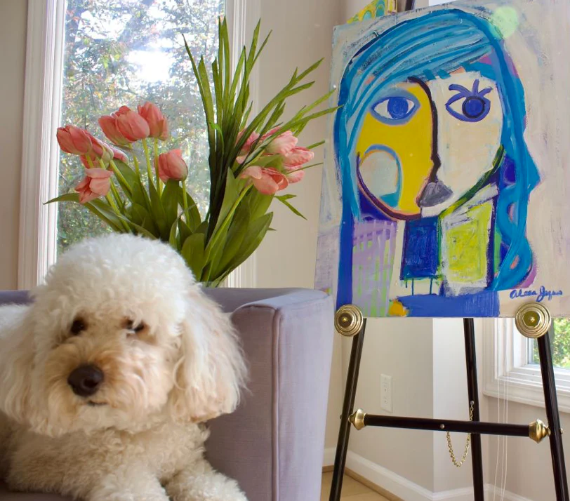 A blue abstract portrait painting hangs in a contemporary living room with a dog sitting in the foreground.