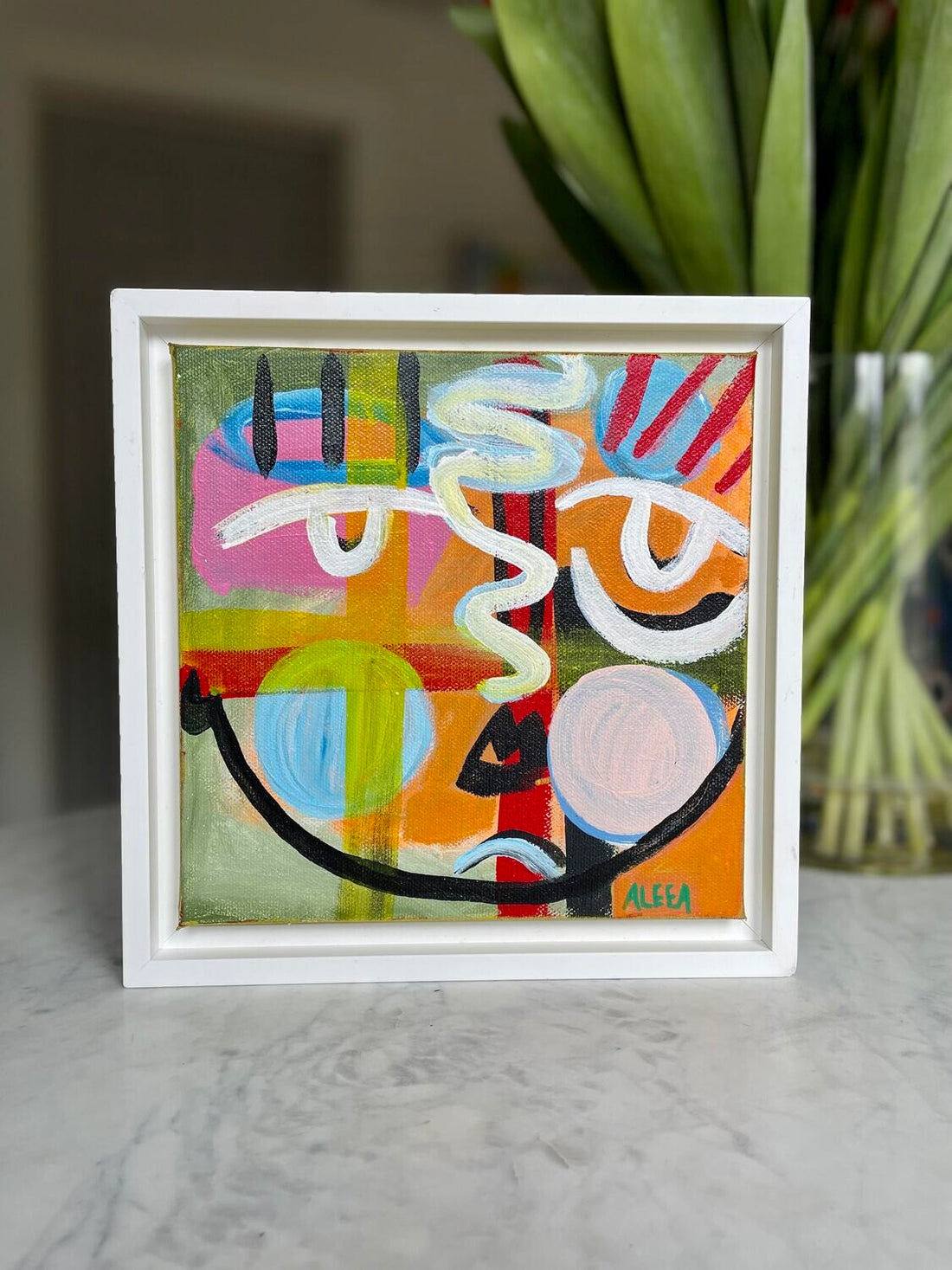 A framed colorful abstract portrait sits on a marble table in front of a vase of flowers.